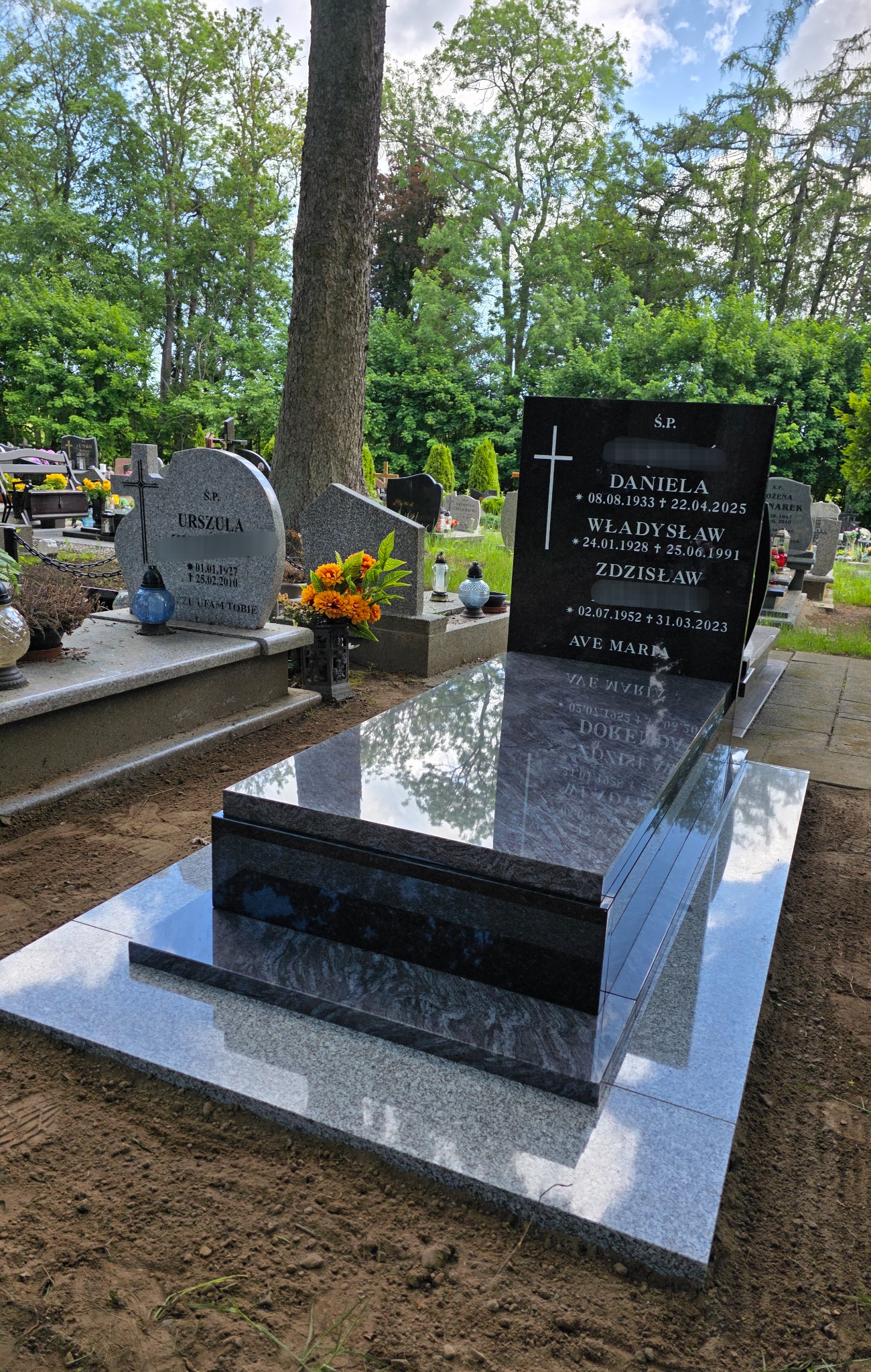 Polerowany, grafitowy nagrobek z czarną tablicą inskrypcyjną na cmentarzu, otoczony zielenią. Widoczne inne groby w tle, drzewo i kwiaty.