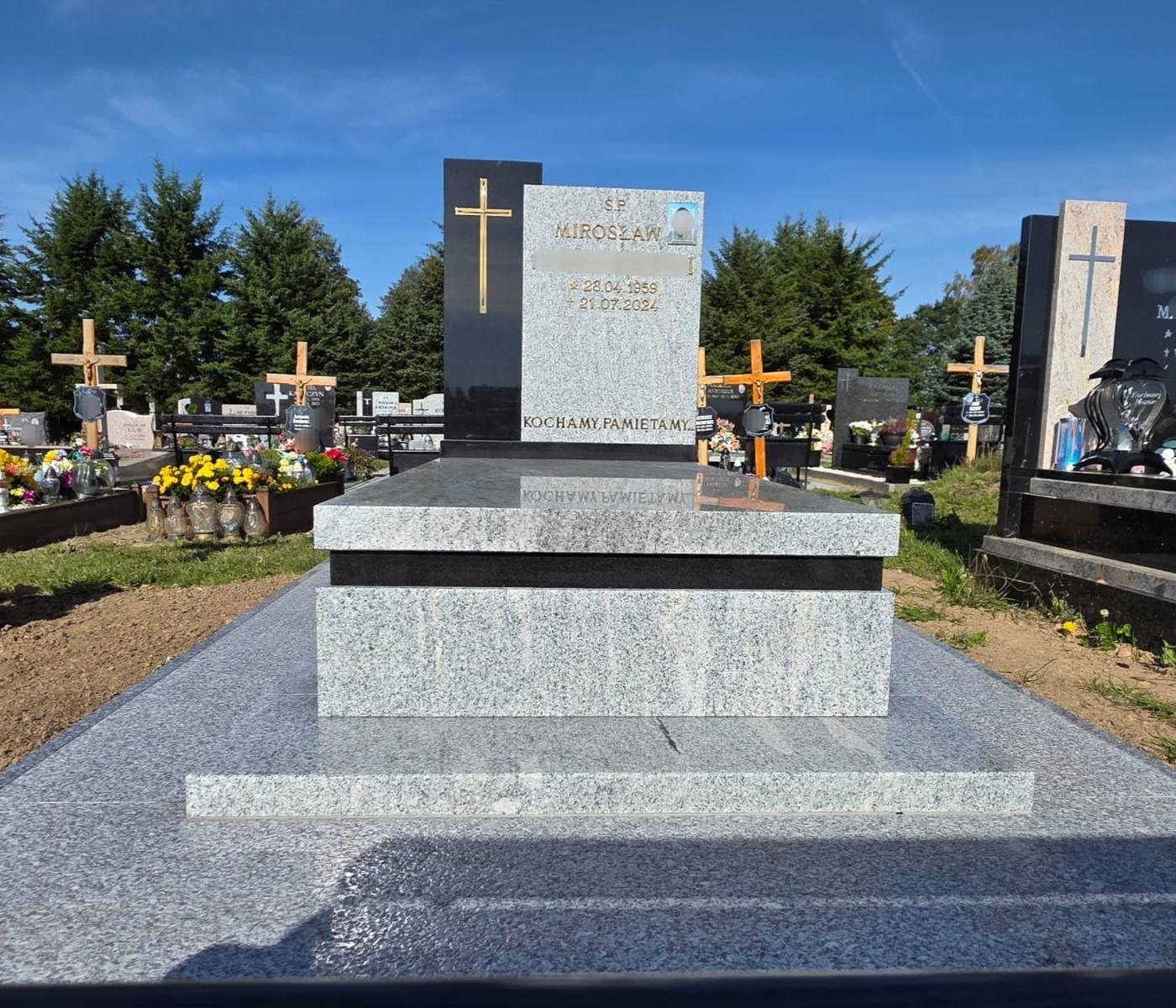 Granitowy nagrobek z czarnym akcentem i złotym krzyżem na cmentarzu, otoczony zielenią drzew. Widoczny portret zmarłego i inskrypcje.