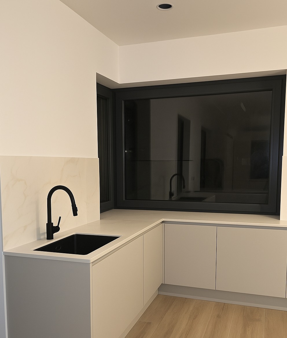 Jasny blat kamienny z czarnym zlewem i baterią, zamontowany w nowoczesnej kuchni z oknem. Minimalistyczny design, neutralna kolorystyka, drewniana podłoga.