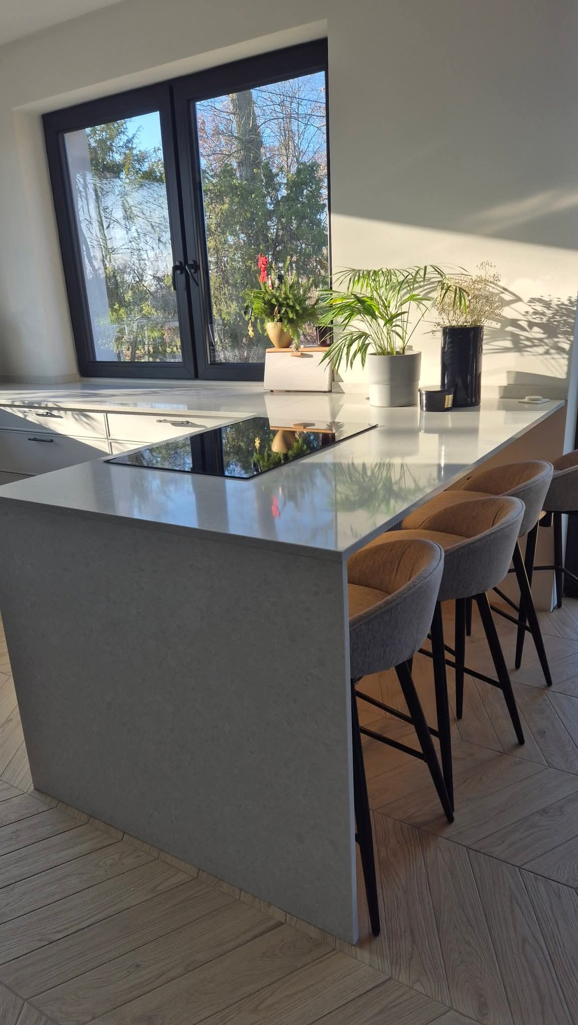 Jasny blat kuchenny z konglomeratu z wbudowaną płytą indukcyjną, oświetlony naturalnym światłem, z trzema krzesłami barowymi i parkietem w jodełkę.