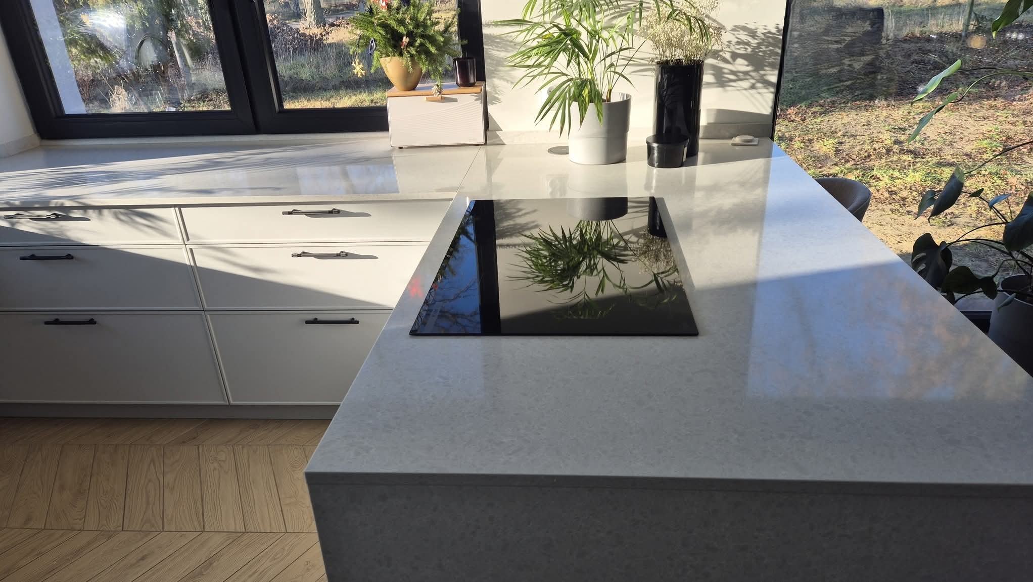 Jasny blat kamienny w kuchni z płytą indukcyjną, odbijającą rośliny i okno. Minimalistyczny design, naturalne światło, drewniana podłoga w jodełkę.
