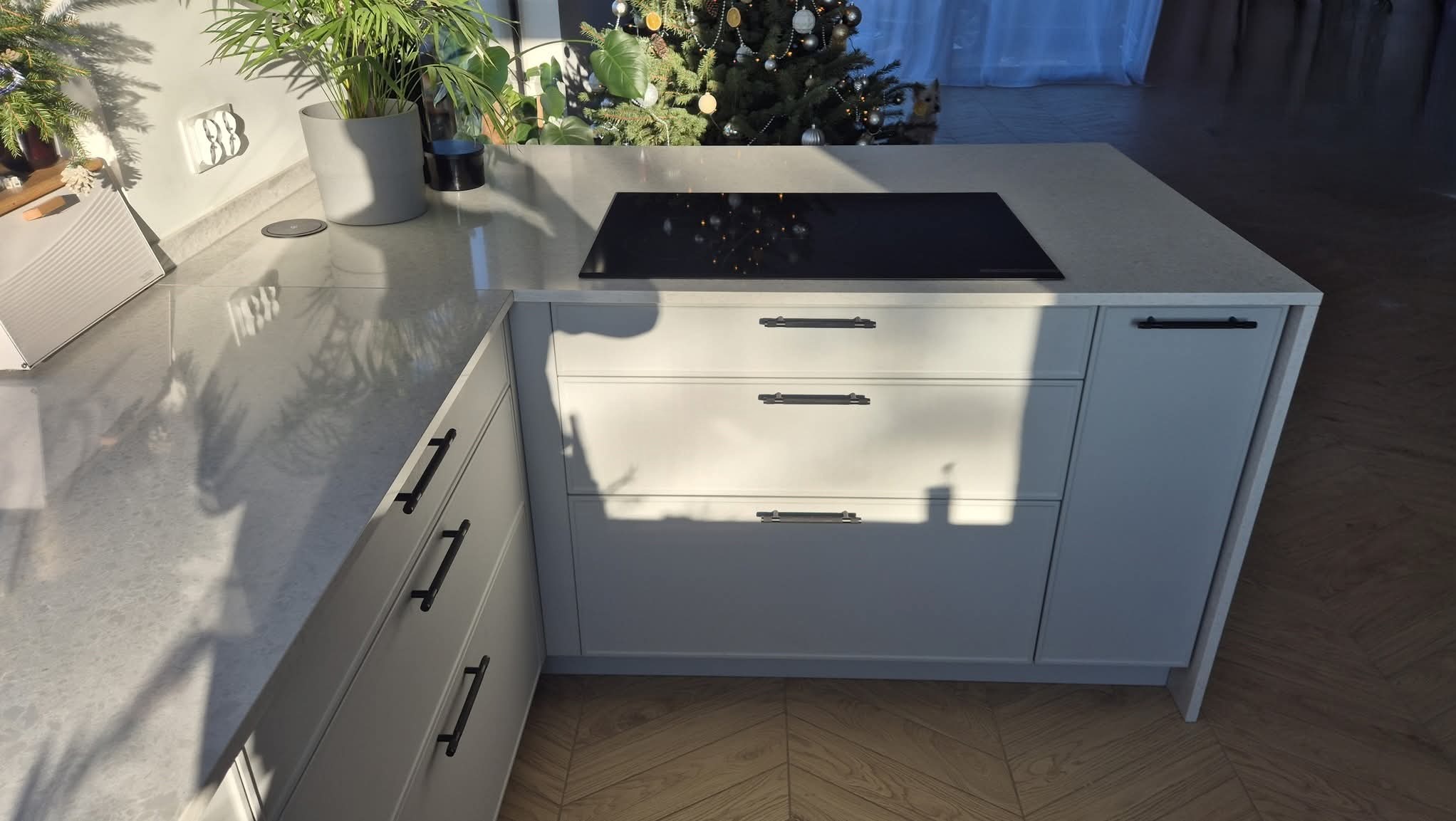 Jasny blat kamienny z wbudowaną płytą indukcyjną na wyspie kuchennej. Widoczne szafki z ciemnymi uchwytami, parkiet w jodełkę i roślina doniczkowa w tle.