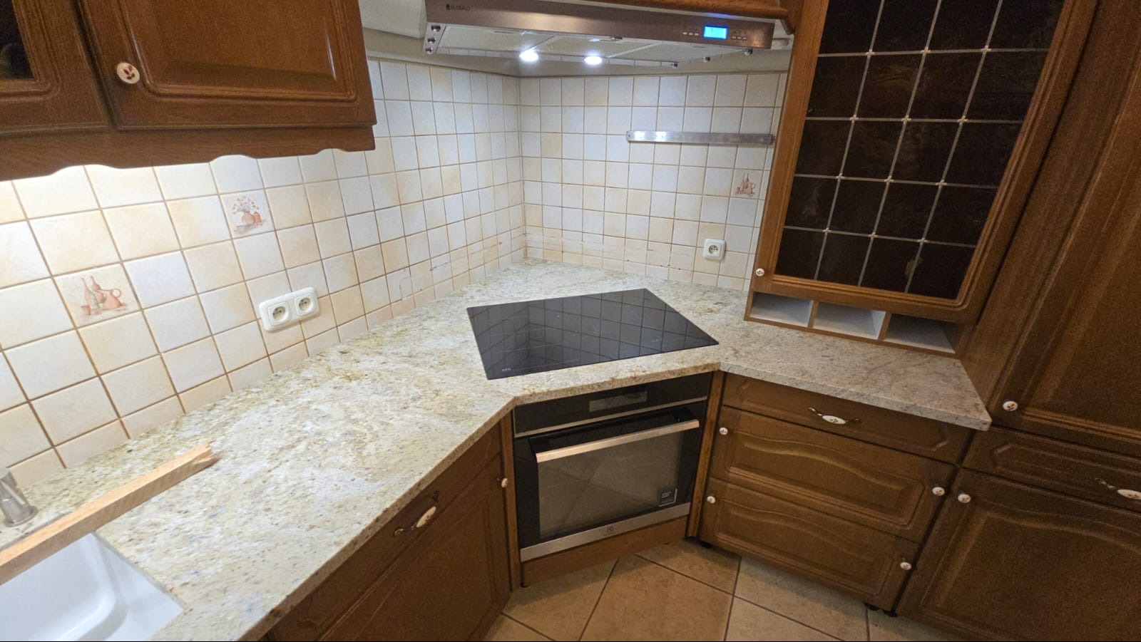 Kuchnia z blatem kamiennym w odcieniach beżu i brązu, z płytą indukcyjną i piekarnikiem. Widoczne drewniane szafki i ceramiczne płytki na ścianie z dekoracyjnymi motywami.