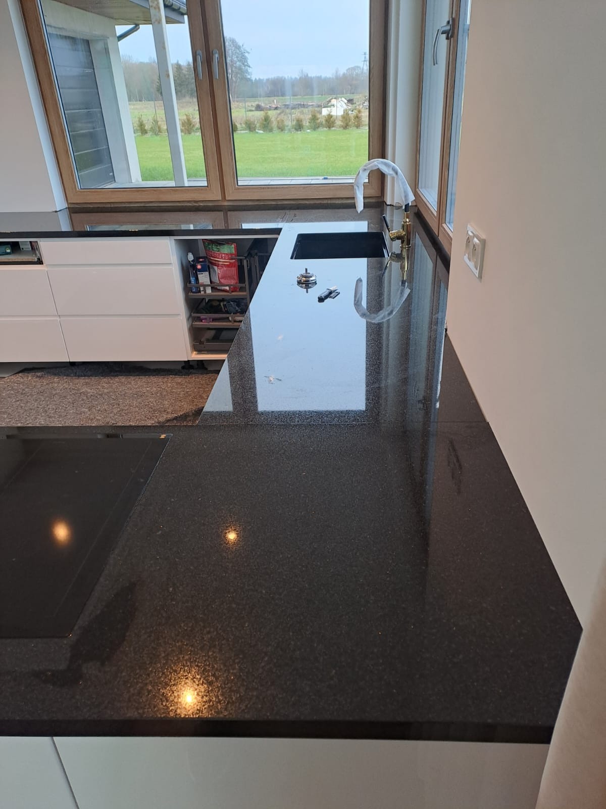 Błyszczący, czarny blat kamienny w kuchni z widokiem na zewnątrz przez okno. Widoczny zlew, bateria i fragment płyty indukcyjnej. Otwarta szafka z akcesoriami kuchennymi.