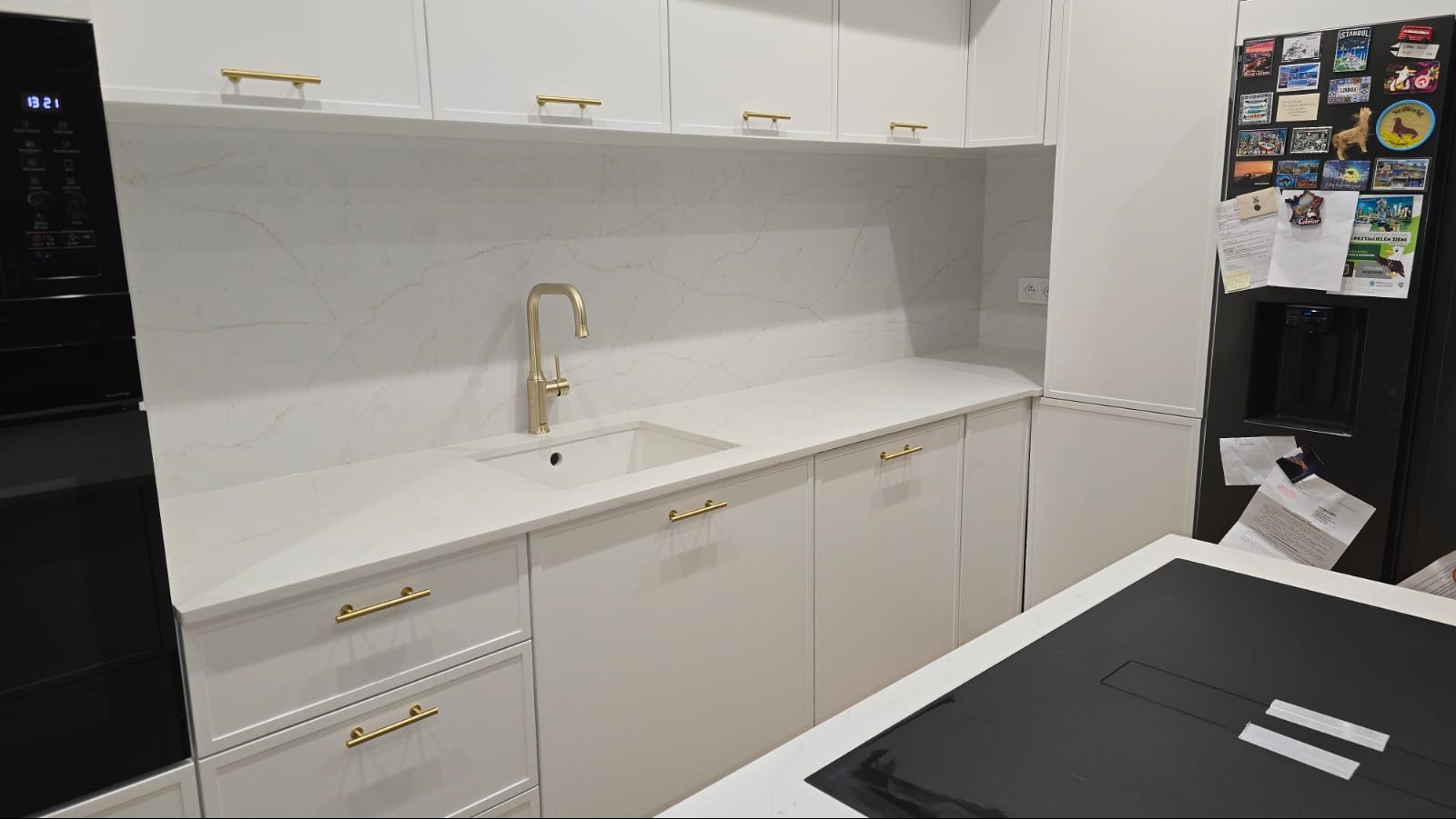 Biała kuchnia z blatem kamiennym o marmurkowym wzorze, złota bateria i uchwyty, czarny piekarnik i lodówka z magnesami. Nowoczesny design.