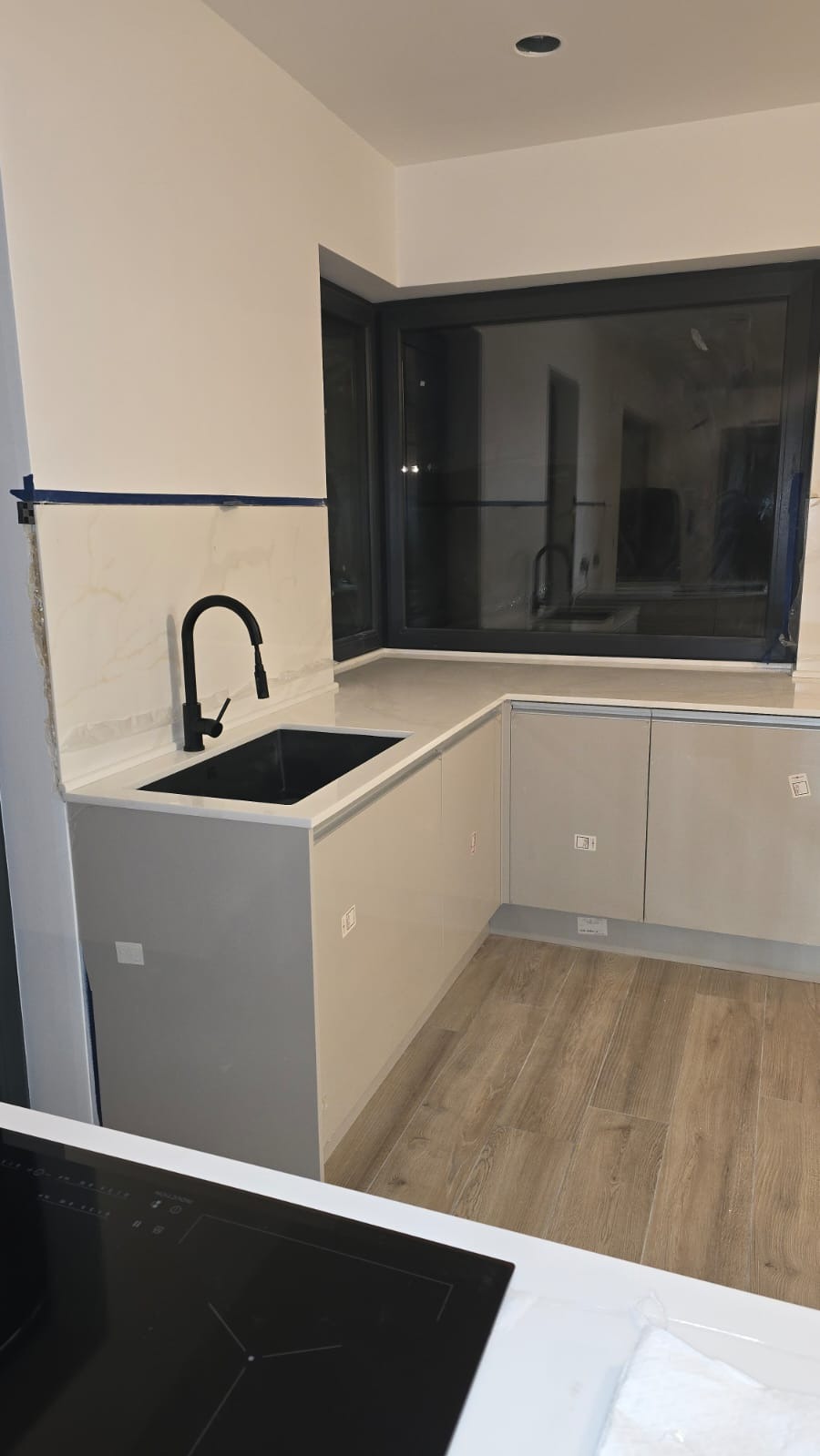 Jasny blat kamienny z czarnym zlewem i baterią w nowoczesnej kuchni z oknem. Widoczna płyta indukcyjna i drewnopodobna podłoga. Minimalistyczny design.