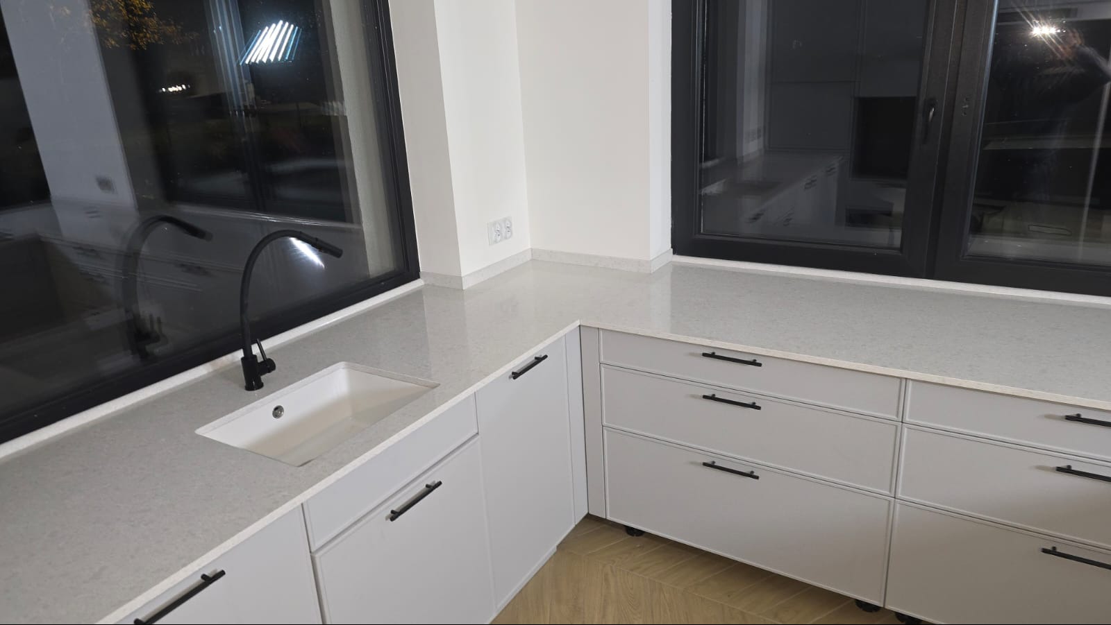 Jasny blat kamienny z umywalką i czarną baterią, zamontowany w nowoczesnej kuchni z szarymi szafkami i dużymi oknami. Minimalistyczny design, neutralna kolorystyka.