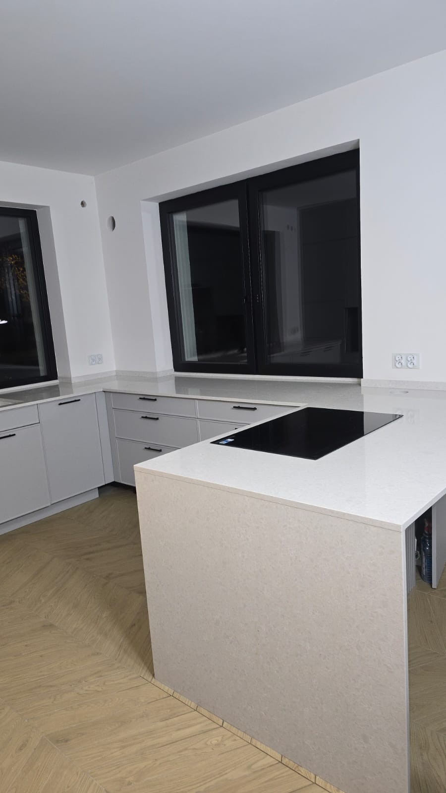 Jasny blat kamienny z wbudowaną płytą indukcyjną w nowoczesnej kuchni z dużym oknem. Parkiet ułożony w jodełkę. Minimalistyczny design.