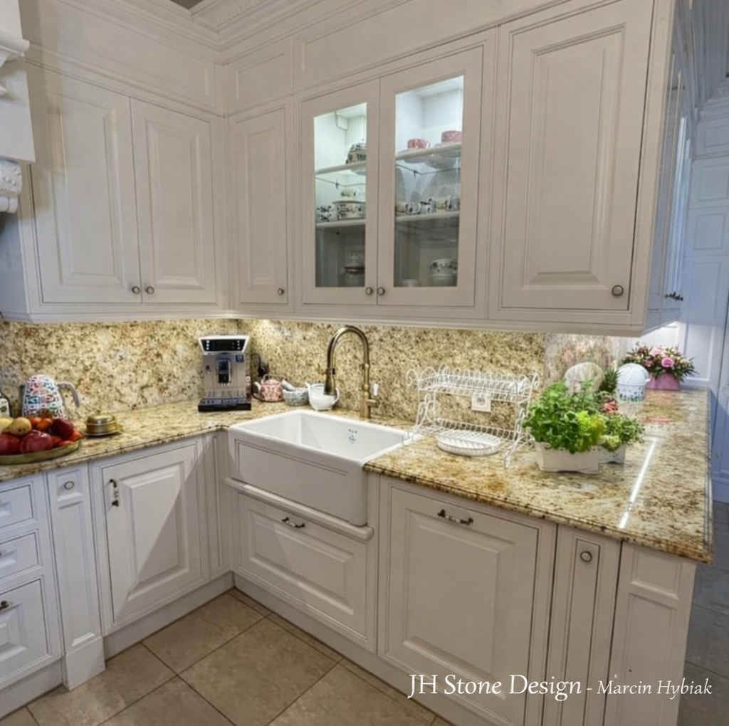 Elegancka kuchnia z blatem z kamienia w odcieniach beżu i brązu, białe szafki z przeszklonymi witrynami, rustykalny zlew i złota armatura. Ciepłe oświetlenie.