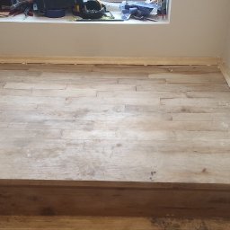 Mikołaj Skrzypczyk - Fragment pokoju ze schodami i jasną, drewnianą podłogą w trakcie układania, widoczny odkurzacz przemysłowy Karcher, listwy przypodłogowe częściowo zamontowane, okno z narzędziami w tle.