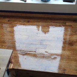 Mikołaj Skrzypczyk - Świeżo polakierowana, błyszcząca podłoga drewniana z zabezpieczonymi listwami przypodłogowymi niebieską taśmą malarską, widoczny fragment drewnianych schodów.