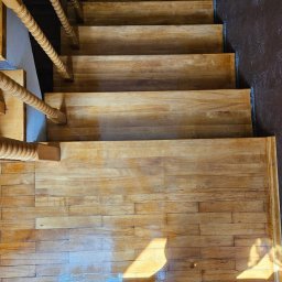 Mikołaj Skrzypczyk - Widok z góry na odnowione drewniane schody z ozdobną balustradą i fragmentem parkietu na podłodze, na których widoczny jest refleks światła.