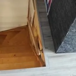 Widok na schody drewniane z poziomu podłogi wyłożonej panelami w odcieniach szarości, fragment regału z książkami w tle.