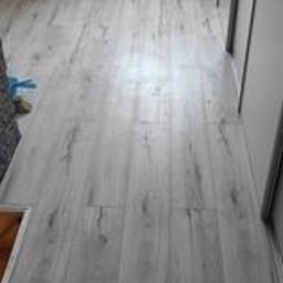 Mikołaj Skrzypczyk - Jasnoszare panele podłogowe z widoczną strukturą drewna, ułożone wzdłużnie w korytarzu, z fragmentem regału z książkami w tle po lewej stronie.