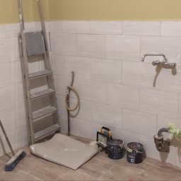 Mikołaj Skrzypczyk - Wnętrze łazienki w trakcie prac remontowych: białe płytki na ścianach, aluminiowa drabina, zabezpieczona podłoga folią, pędzle, wiaderka z farbą, grzejnik oraz prowizoryczne zabezpieczenia na rurach.