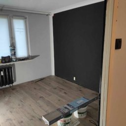 Mikołaj Skrzypczyk - Pokój po remoncie z nową drewnianą podłogą, pomalowaną na czarno ścianą oraz widocznymi puszkami farby i panelami podłogowymi.