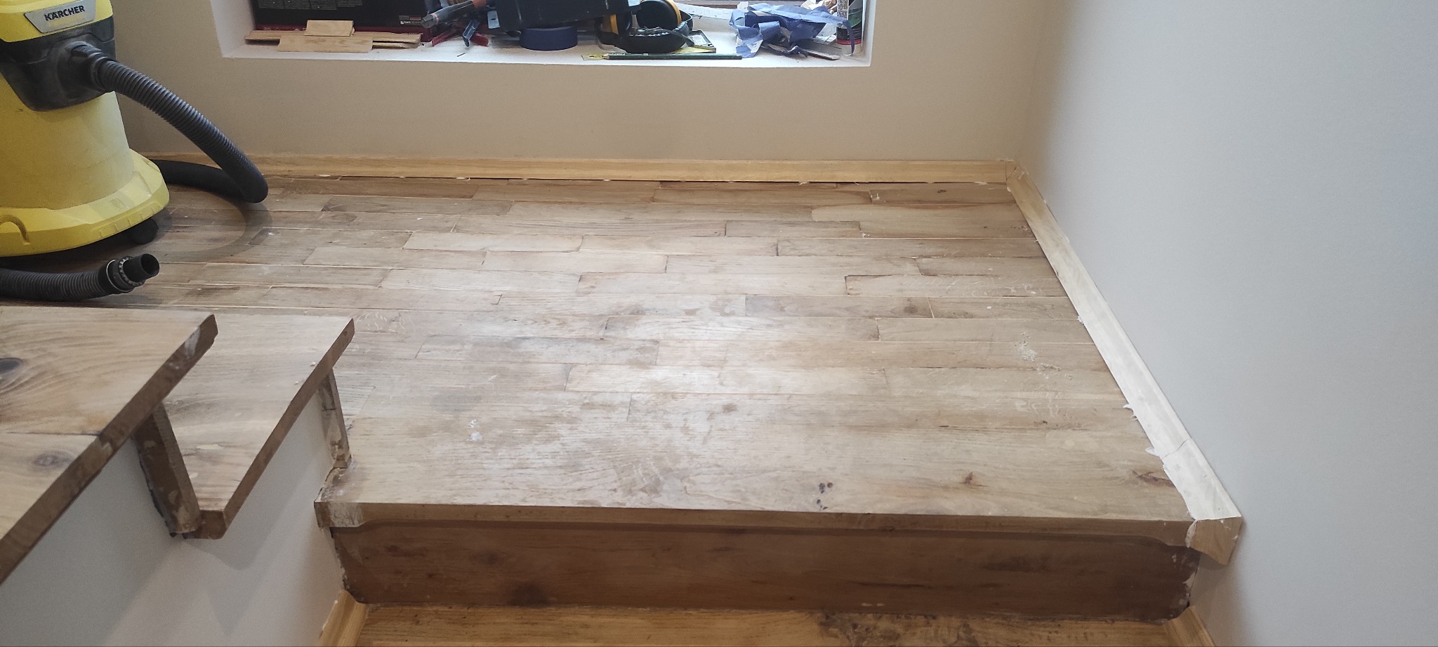 Fragment pokoju ze schodami i jasną, drewnianą podłogą w trakcie układania, widoczny odkurzacz przemysłowy Karcher, listwy przypodłogowe częściowo zamontowane, okno z narzędziami w tle.