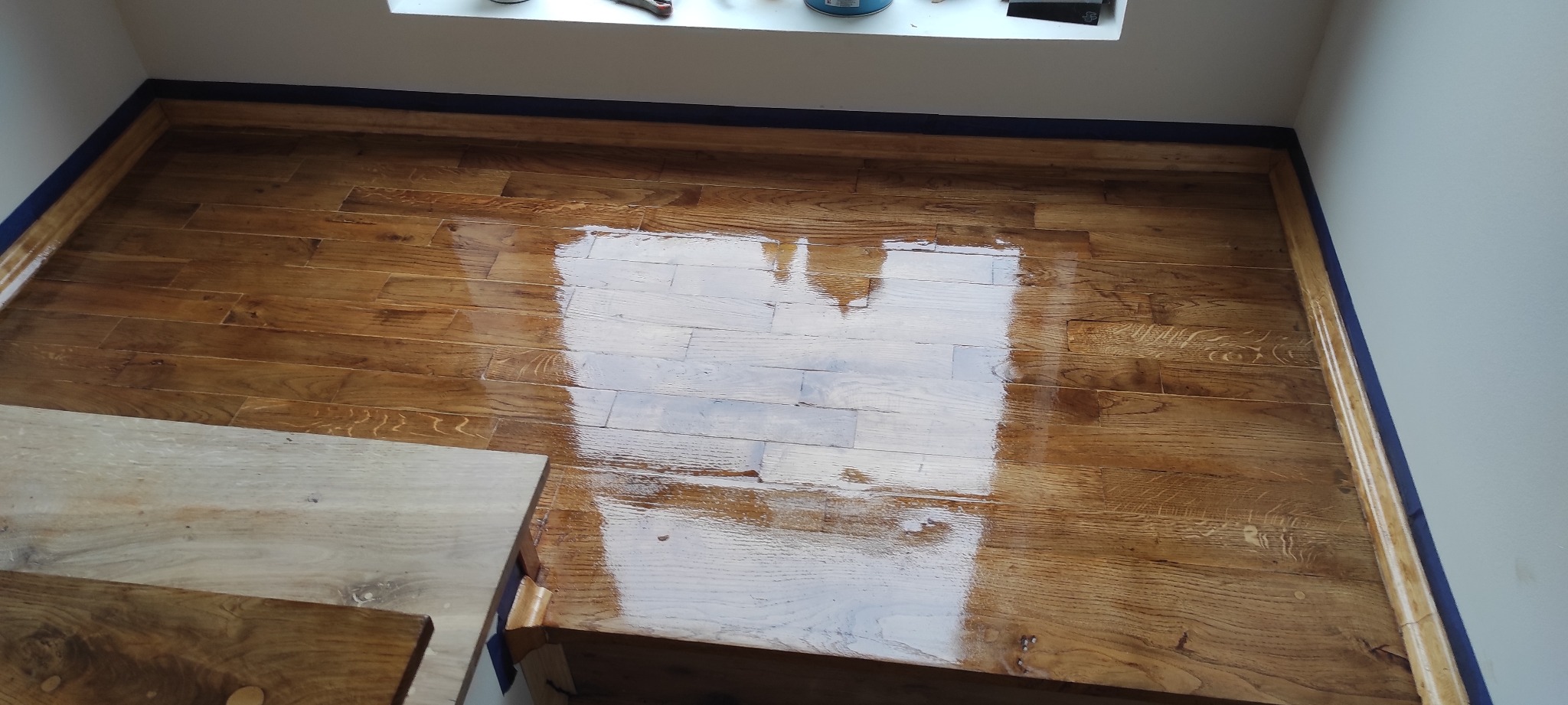 Świeżo polakierowana, błyszcząca podłoga drewniana z zabezpieczonymi listwami przypodłogowymi niebieską taśmą malarską, widoczny fragment drewnianych schodów.