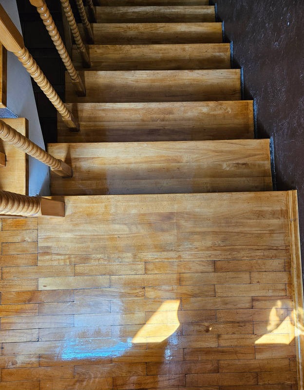 Widok z góry na odnowione drewniane schody z ozdobną balustradą i fragmentem parkietu na podłodze, na których widoczny jest refleks światła.