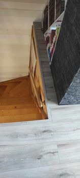 Widok na schody drewniane z poziomu podłogi wyłożonej panelami w odcieniach szarości, fragment regału z książkami w tle.