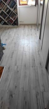 Jasnoszare panele podłogowe z widoczną strukturą drewna, ułożone wzdłużnie w korytarzu, z fragmentem regału z książkami w tle po lewej stronie.