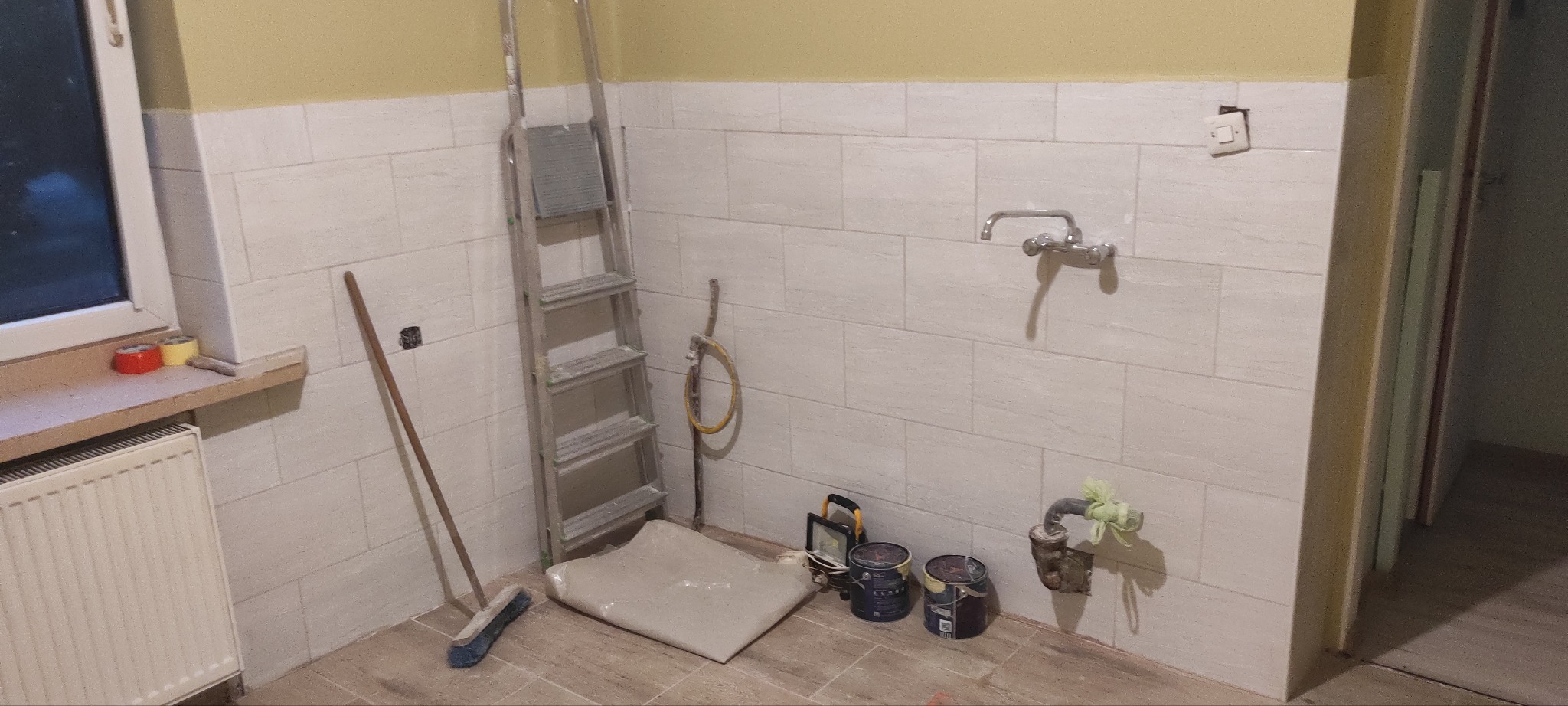Wnętrze łazienki w trakcie prac remontowych: białe płytki na ścianach, aluminiowa drabina, zabezpieczona podłoga folią, pędzle, wiaderka z farbą, grzejnik oraz prowizoryczne zabezpieczenia na rurach.
