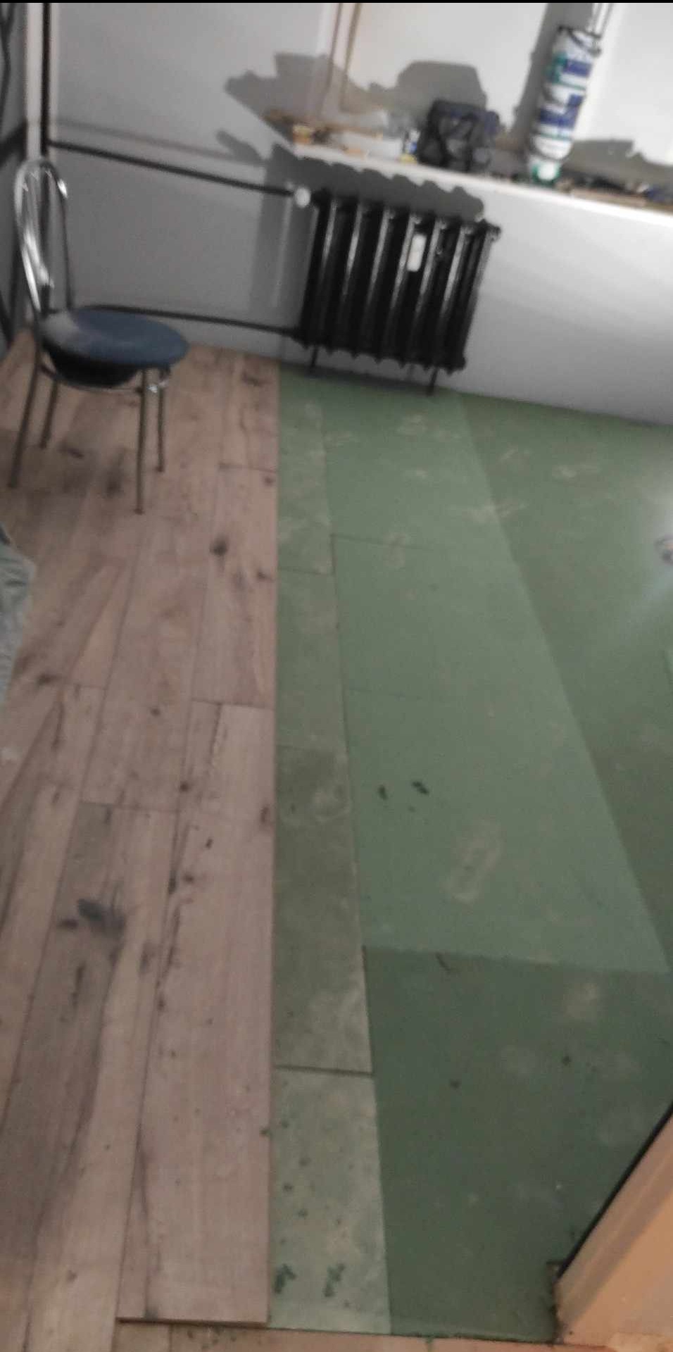 Fragment podłogi w trakcie prac: część wyłożona panelami imitującymi drewno, część z zielonym podkładem, w tle grzejnik i krzesło.