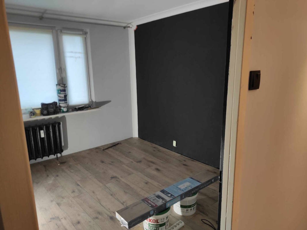 Pokój po remoncie z nową drewnianą podłogą, pomalowaną na czarno ścianą oraz widocznymi puszkami farby i panelami podłogowymi.
