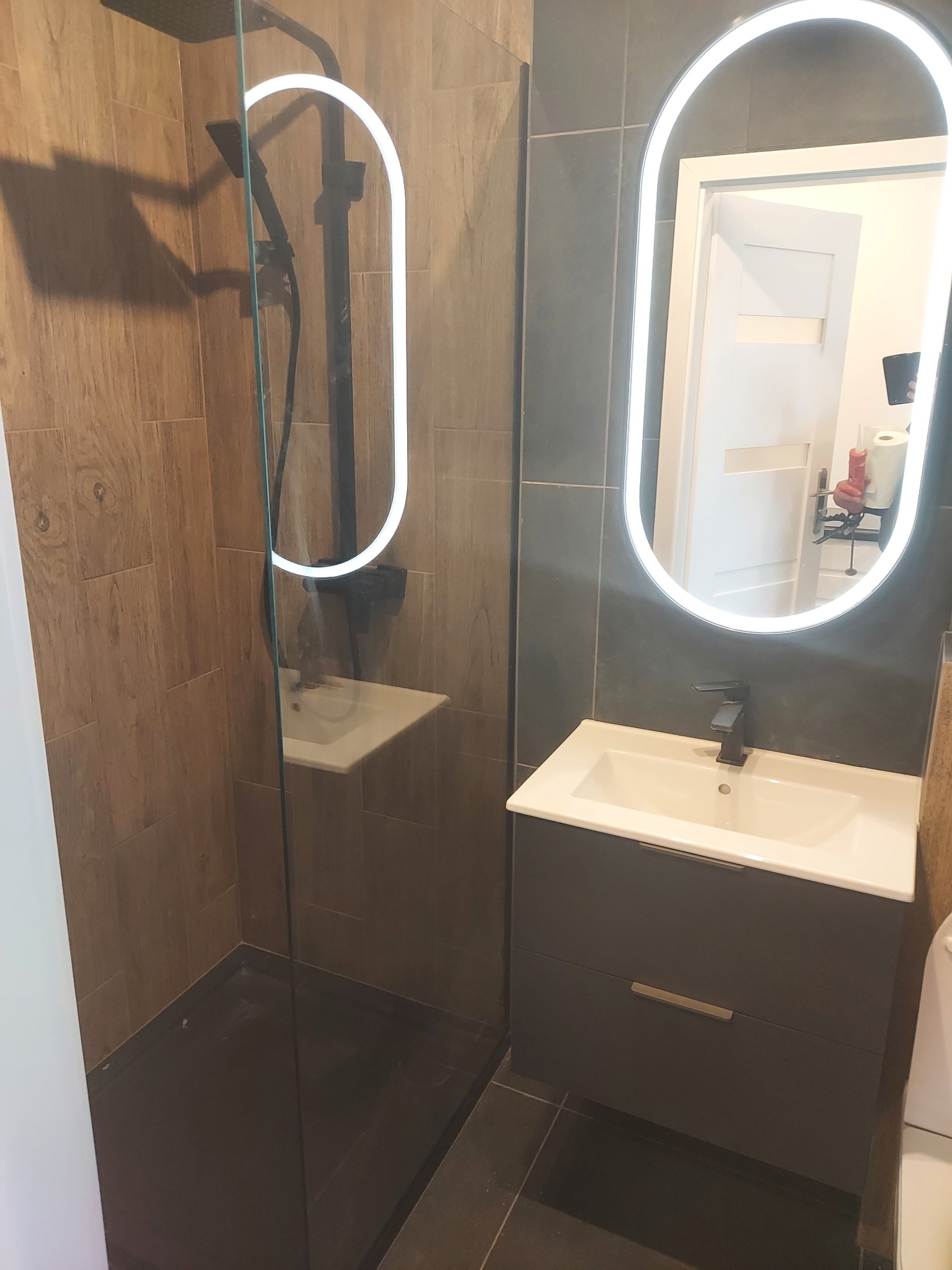 Nowoczesna łazienka z kabiną prysznicową typu walk-in, umywalką z szafką w kolorze grafitowym i podświetlanym lustrem o owalnym kształcie, wykończona płytkami w odcieniach brązu i szarości.