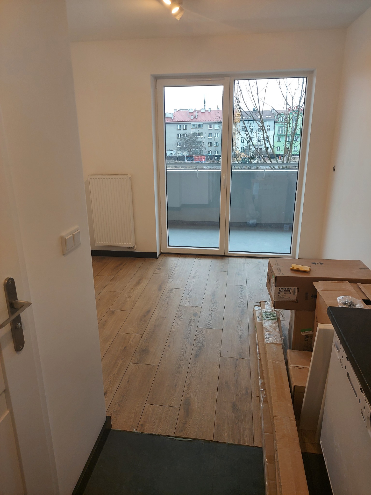 Puste mieszkanie po remoncie z drewnianą podłogą, widoczne kartony z wyposażeniem i balkon z widokiem na budynki.