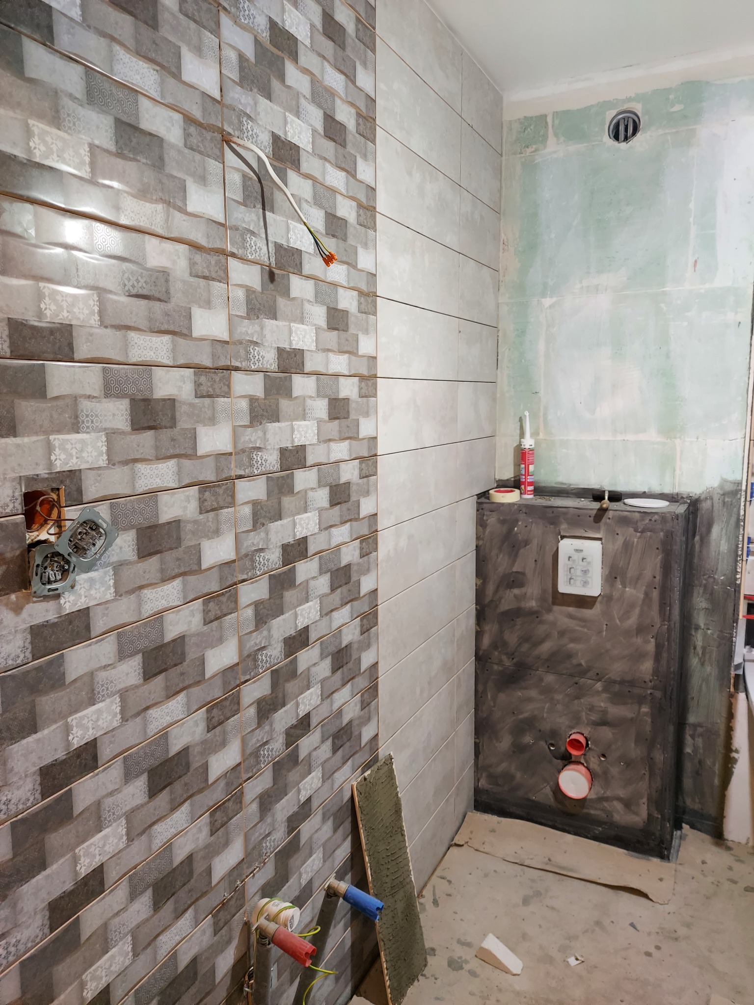 Ściana łazienki w trakcie układania płytek w odcieniach szarości, widoczne wystające kable elektryczne i rury hydrauliczne, przygotowana zabudowa pod stelaż WC z uszczelnieniem hydroizolacyjnym.