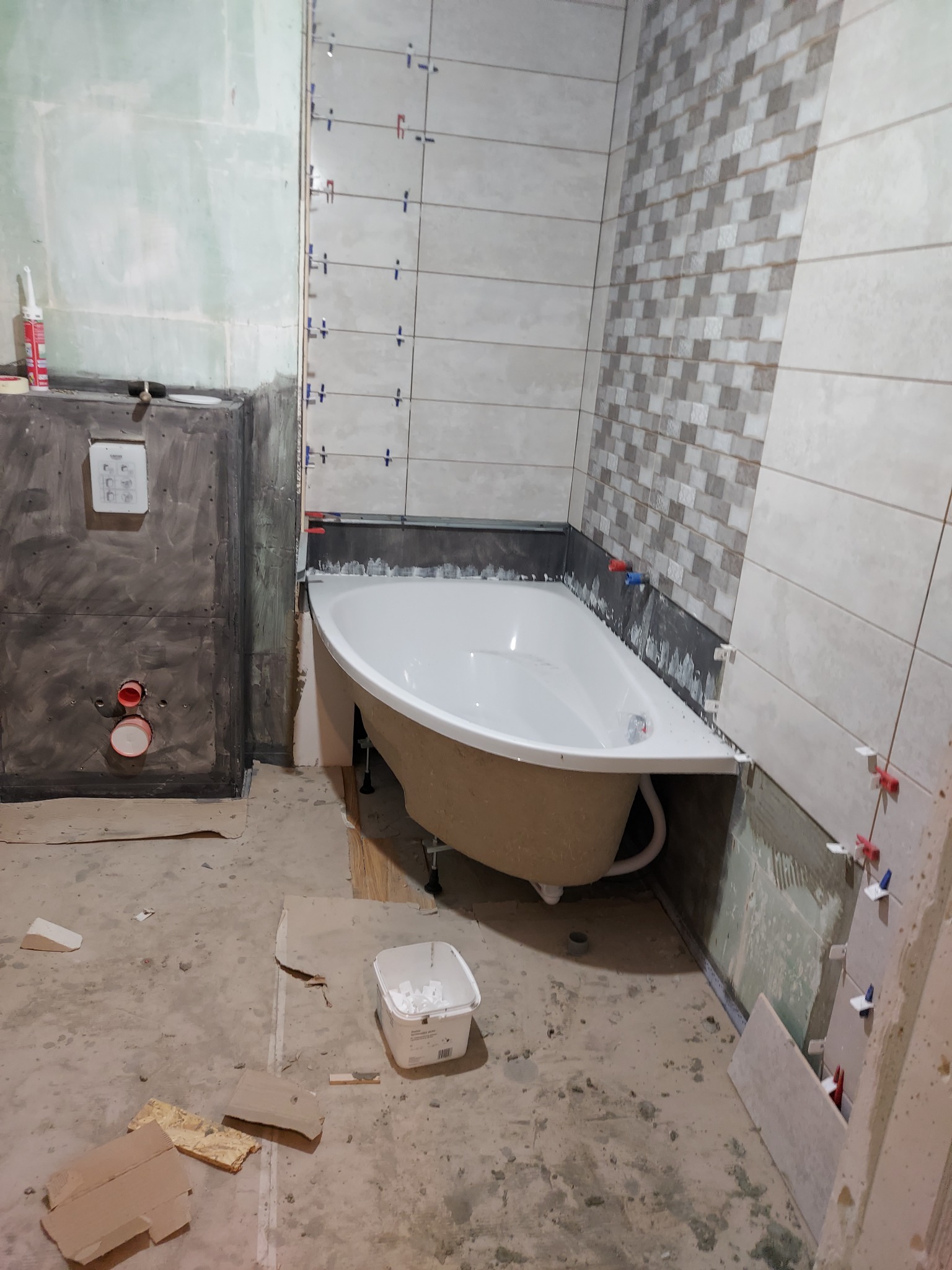 Montaż płytek w łazience: widoczna wanna obudowana płytami, częściowo wyłożona płytkami w dwóch wzorach, z krzyżykami dystansowymi, surowy beton na podłodze i ścianach w trakcie prac wykończeniowych.