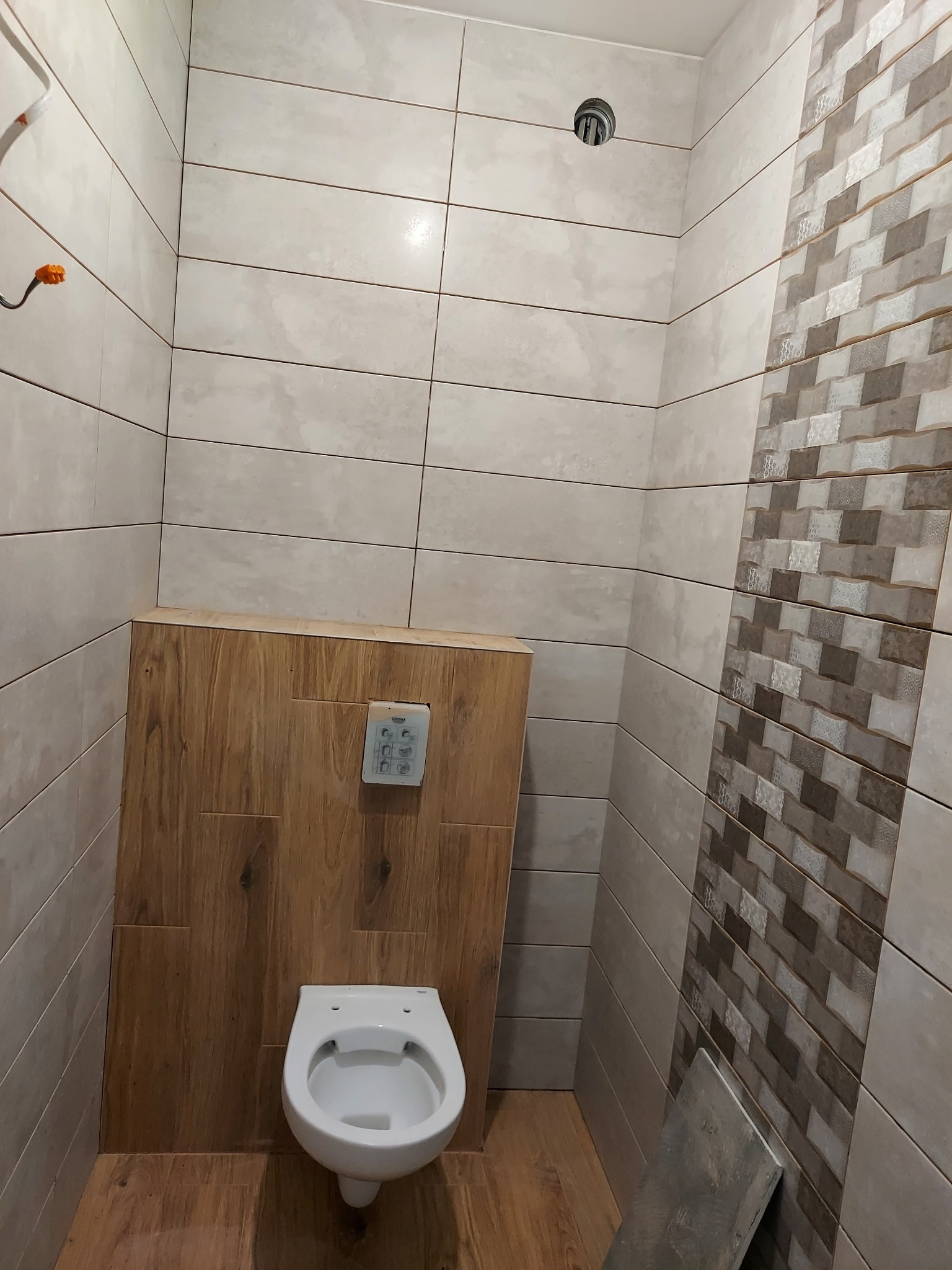 Wnętrze toalety w trakcie remontu, z częściowo ułożonymi płytkami w odcieniach beżu i brązu, widoczna podtynkowa instalacja spłuczki i wisząca muszla klozetowa bez deski.