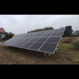 Panel fotowoltaiczny zamontowany na metalowej konstrukcji, widoczny na tle pochmurnego nieba i drzew w oddali.