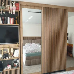 Szafa z przesuwnymi drzwiami lustrzanymi z drewnianymi listwami, wbudowana w ścianę, z półkami z boku, na których stoją książki i dekoracje, z widokiem na sypialnię z łóżkiem.