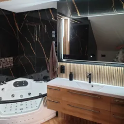 Nowoczesna łazienka z narożną wanną z hydromasażem, drewnianą szafką pod umywalką i podświetlanym lustrem na tle czarnych płytek z złotymi żyłami.