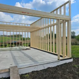 Wznoszenie drewnianej konstrukcji szkieletowej budynku na betonowej płycie fundamentowej. Widoczne stemple na elementach szkieletu. W tle zieleń łąk i drzew pod błękitnym niebem.