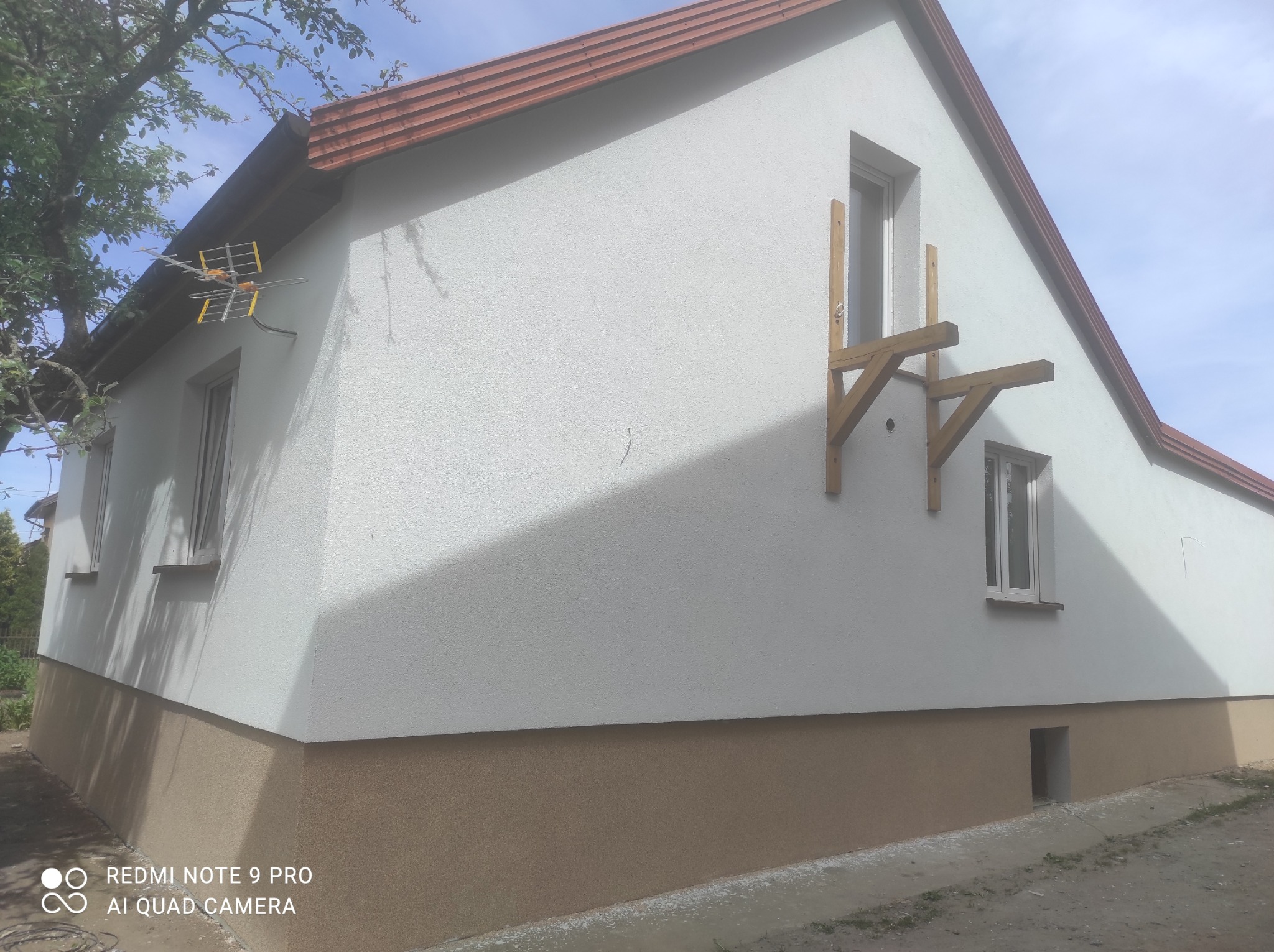 Świeżo pomalowana elewacja domu jednorodzinnego z widocznymi drewnianymi elementami podtrzymującymi okno, dwukolorowa fasada, antena telewizyjna na ścianie bocznej.