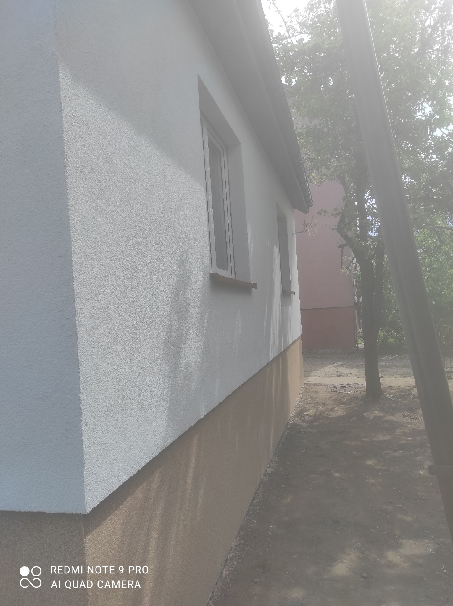Boczny widok na świeżo otynkowany dom z dwoma oknami, dolna część elewacji w kolorze brązowym, górna w kolorze białym, z widocznym drzewem i słupem energetycznym w tle.