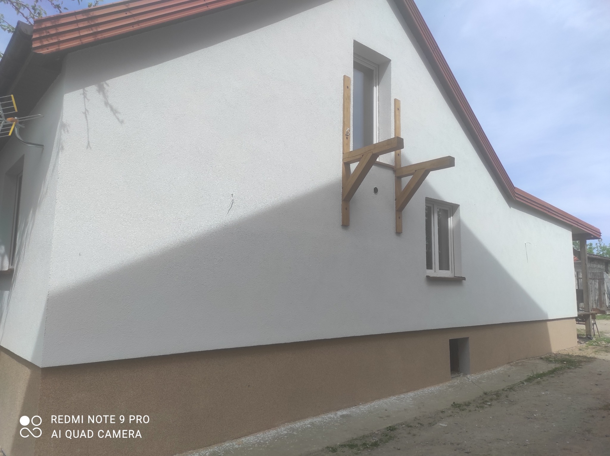 Boczny widok na dom z jasną, teksturowaną elewacją i brązowym cokołem; widoczne drewniane rusztowanie przy oknie na górze i antena na dachu.