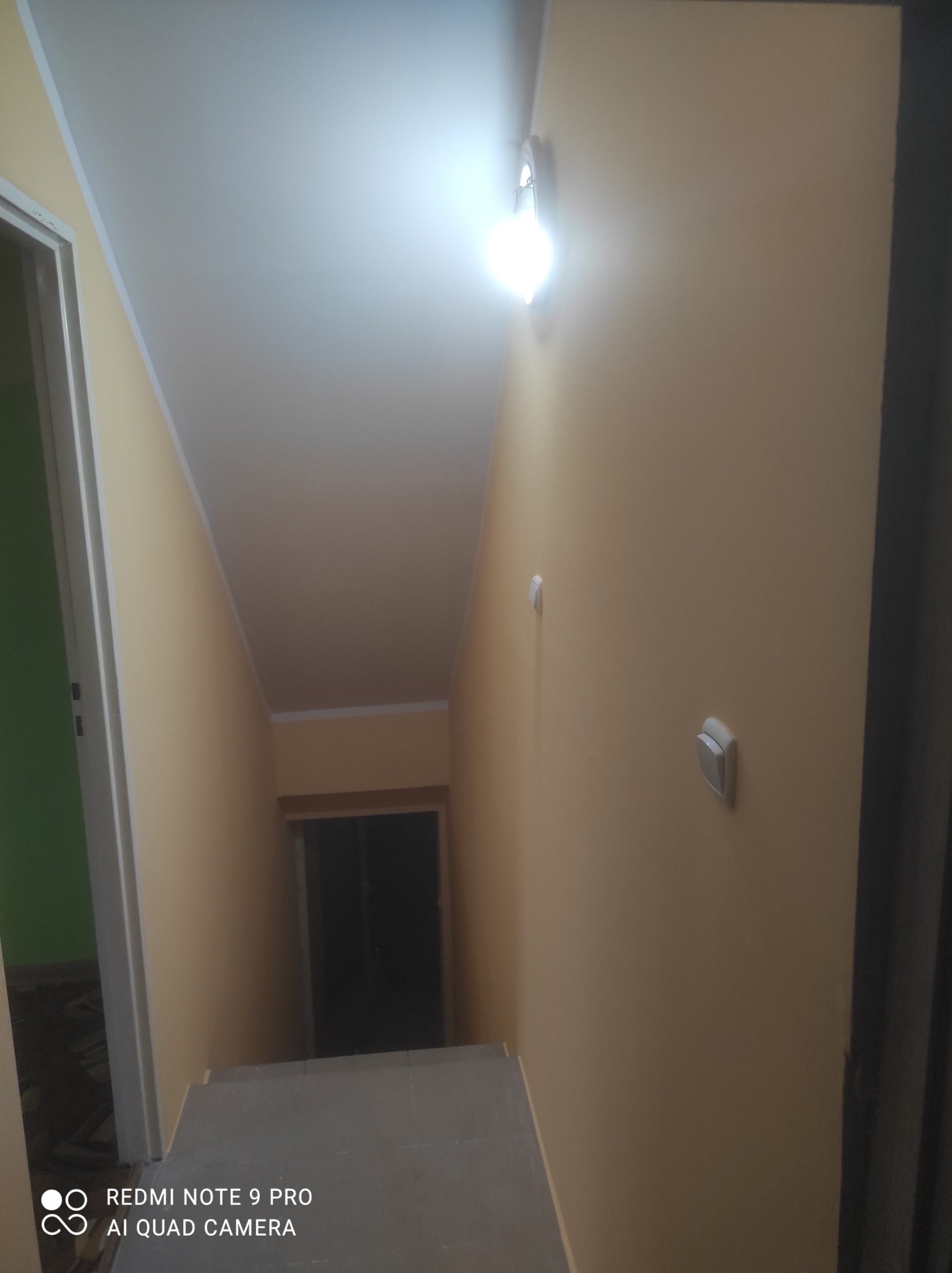 Klatka schodowa z pomalowanymi na beżowo ścianami, widoczne schody z szarego lastryko, biała oprawa oświetleniowa na ścianie i fragment zielonej ściany za otwartymi drzwiami.