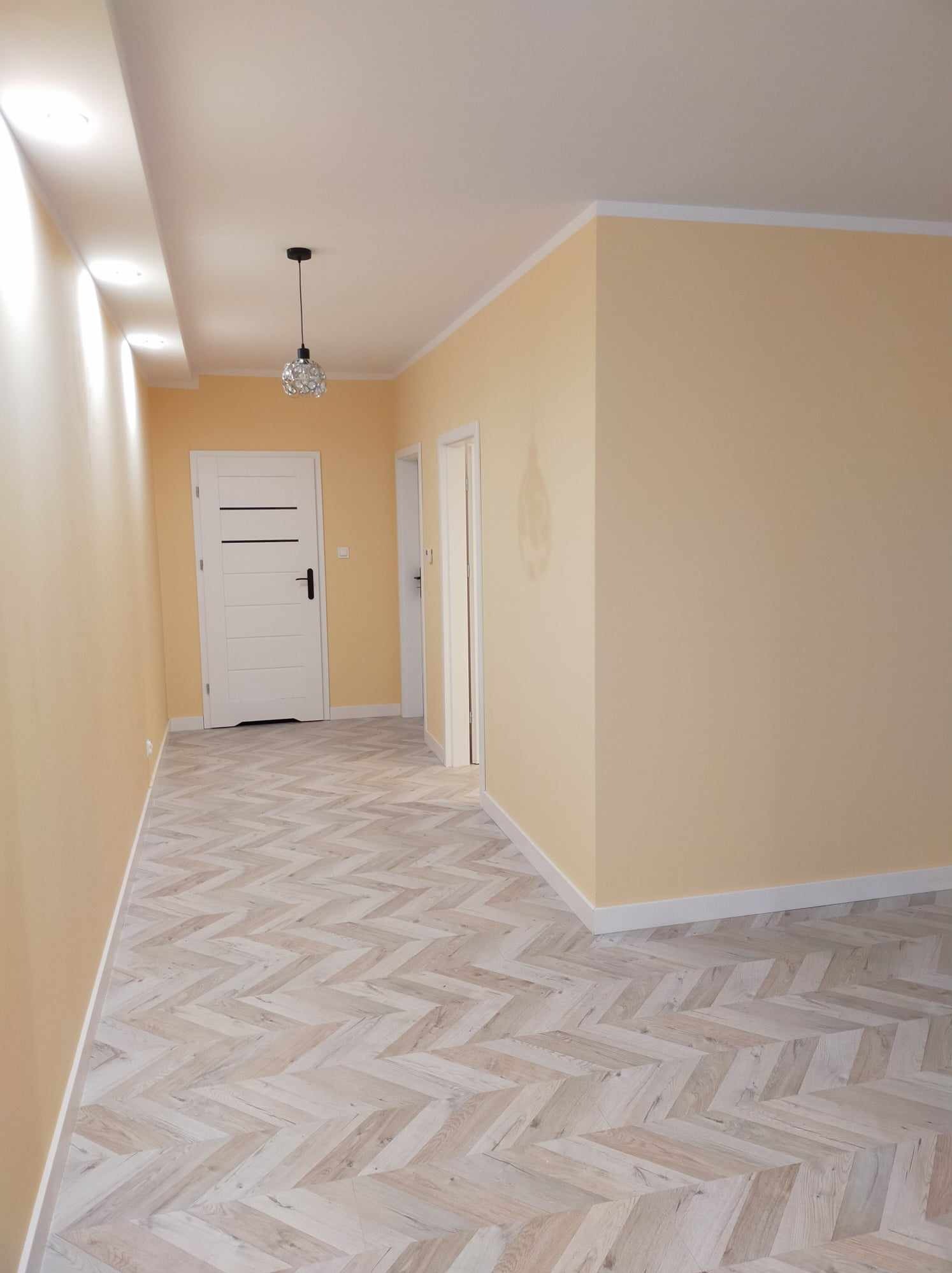 Świeżo pomalowane, jasnożółte ściany w korytarzu w Szydłowie, z nową podłogą w jodełkę i białymi listwami przypodłogowymi oraz ozdobnym żyrandolem.