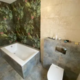 Remont łazienki w Lesznie: biała wanna obudowana szarą mozaiką, toaleta podwieszana oraz ściana z wzorem tropikalnych liści i jasnych płytek, widoczne narzędzia i materiały budowlane.