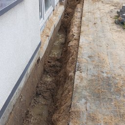 GRZECH-BUD Grzegorz Ludwiczak - Wykop wzdłuż ściany budynku z widoczną warstwą izolacji fundamentowej, częściowo wypełniony wodą i błotem, obok ułożona kostka brukowa.