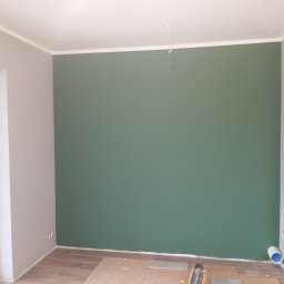 GRZECH-BUD Grzegorz Ludwiczak - Świeżo pomalowane wnętrze z jedną ścianą w odcieniu stonowanej zieleni, kontrastującą z szarą ścianą boczną i jasnym sufitem z widocznym przewodem. Podłoga częściowo wyłożona panelami...