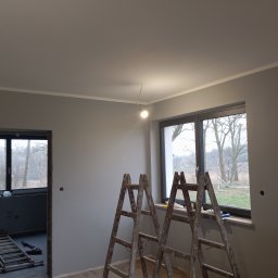 GRZECH-BUD Grzegorz Ludwiczak - Świeżo pomalowane szare ściany z białym wykończeniem, dwie drewniane drabiny opierają się o ścianę pod oknem, w tle widoczne jest pomieszczenie w trakcie remontu.