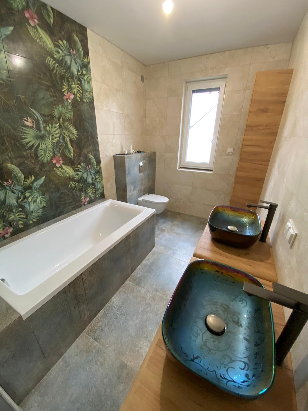Nowoczesna łazienka z wanną, toaletą podtynkową i dwiema szklanymi umywalkami w odcieniach niebieskiego i brązu na drewnianym blacie, ściana z motywem tropikalnych liści.