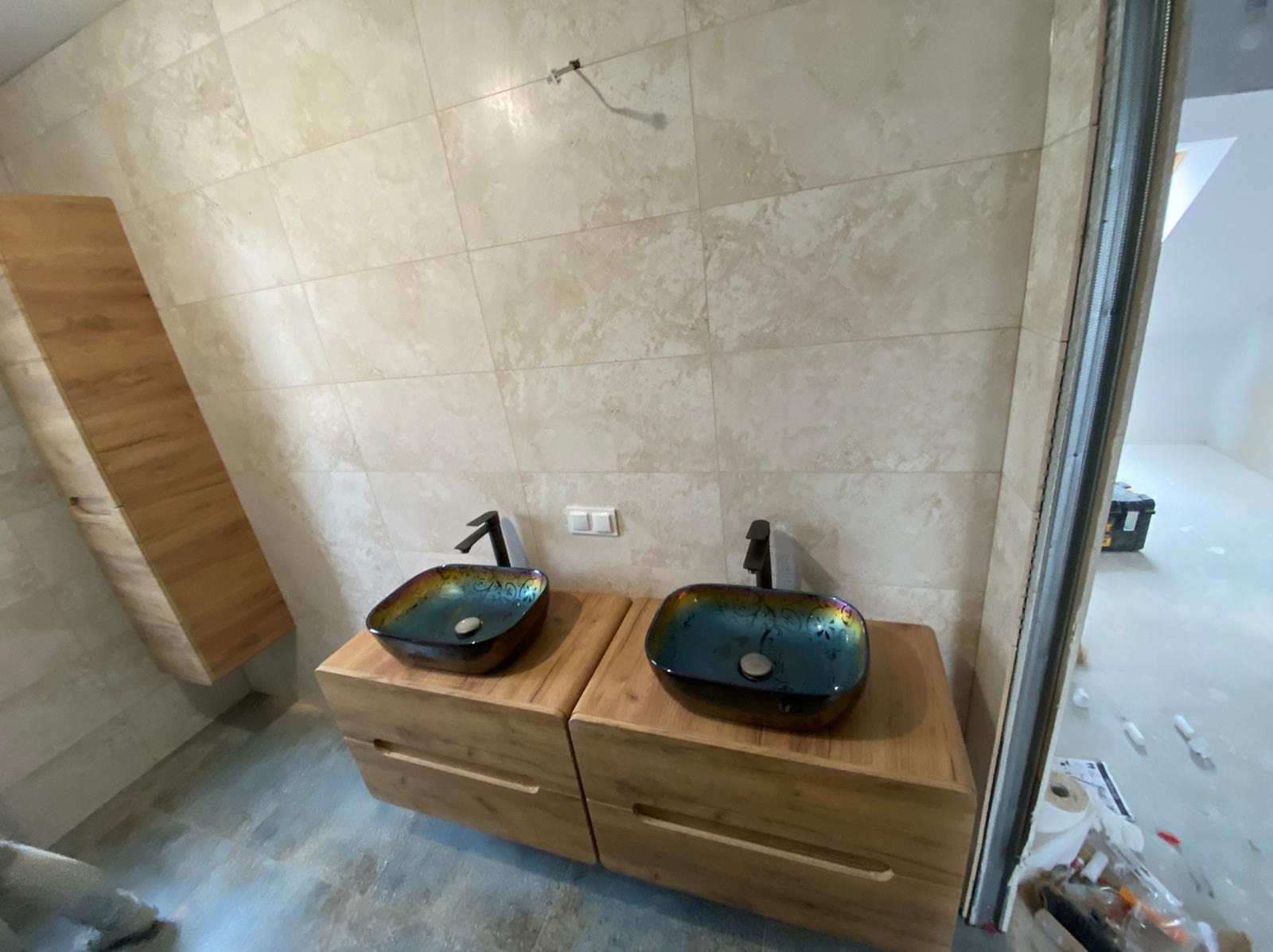 Dwie umywalki z czarnymi bateriami, każda osadzona na drewnianej szafce z szufladami, na tle beżowych płytek ściennych i częściowo widocznego remontowanego pomieszczenia.