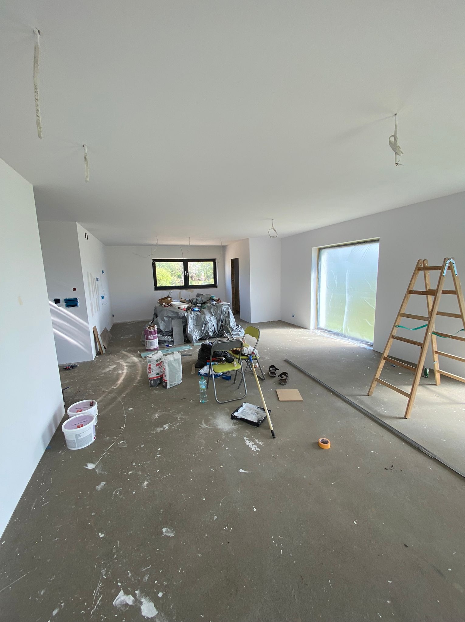 Wnętrze w trakcie prac wykończeniowych: świeżo pomalowane białe ściany, zabezpieczone okno folią, drabina, wiaderka z farbą, narzędzia malarskie i meble przykryte folią ochronną, widoczne ślady...