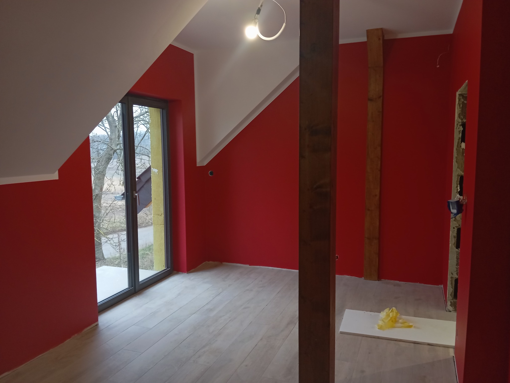 Pokój na poddaszu po remoncie, ściany pomalowane na intensywny czerwony kolor, jasne panele podłogowe, widoczne drewniane elementy konstrukcyjne i okno z widokiem na drzewa.